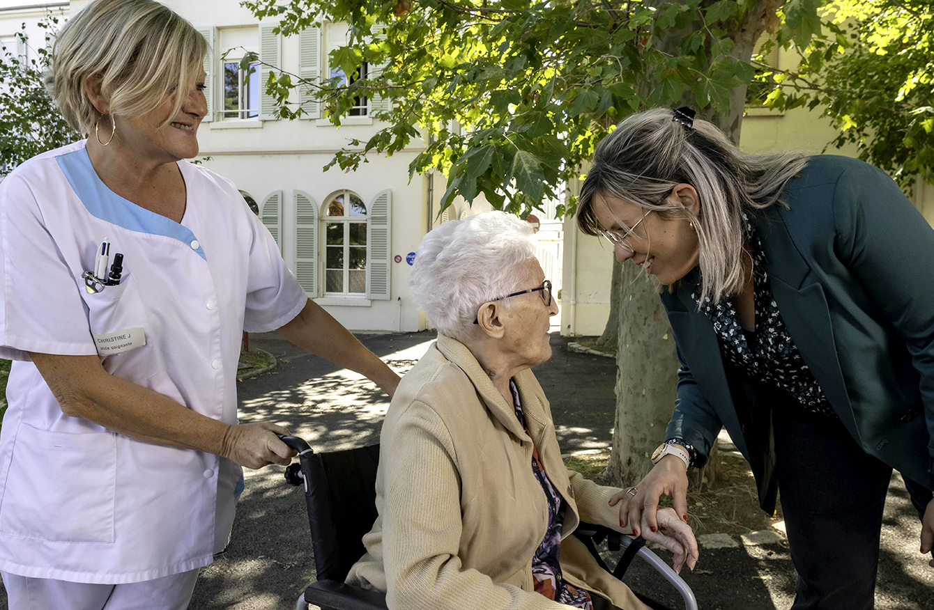 Une dame âgées en fauteuil roulant entourée de deux personnes l'une du corps médical et l'autre du service à la personne.