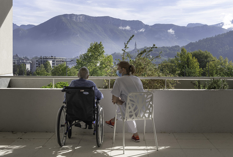Une personne âgée dans un fauteuil roulant et une jeune aide à la personne assise à ses côtés. Tous les deux sont face à un paysage montagneux.